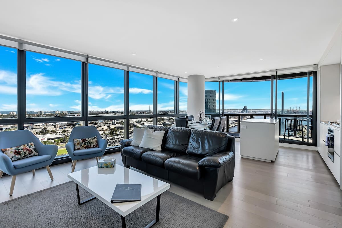 Un salon moderne avec un canapé en cuir noir, deux fauteuils bleus, une table basse blanche et une vue sur la ville grâce à de grandes fenêtres.