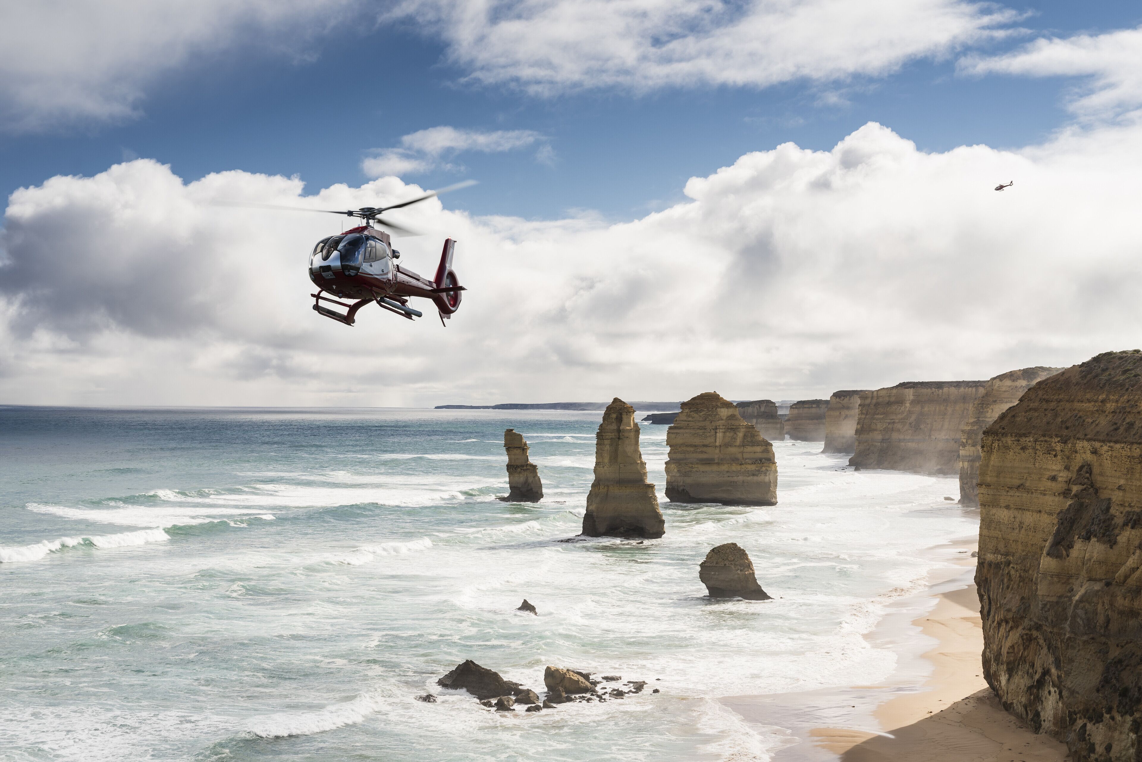 Ein Hubschrauber fliegt über eine Küstenlandschaft mit markanten Felsformationen und einem Strand.