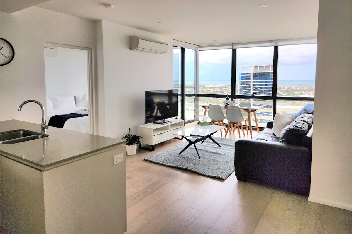 Un appartement moderne comprenant une cuisine, un coin repas et un salon. Il dispose de grandes fenêtres offrant une vue sur la ville, d’un téléviseur à écran plat et d’un canapé.