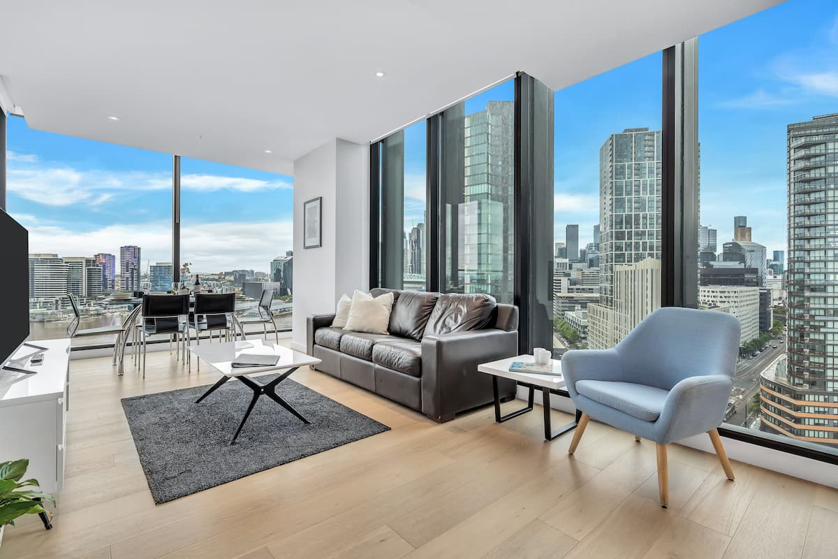 Un salon moderne avec vue sur la ville, comprenant un canapé gris, un fauteuil bleu et un coin repas avec une table et des chaises.