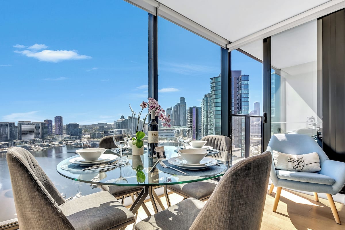 Un espace de restauration moderne avec une table en verre, des chaises recouvertes d’un tissu gris et une vue sur les toits d’une ville et sur un plan d’eau.