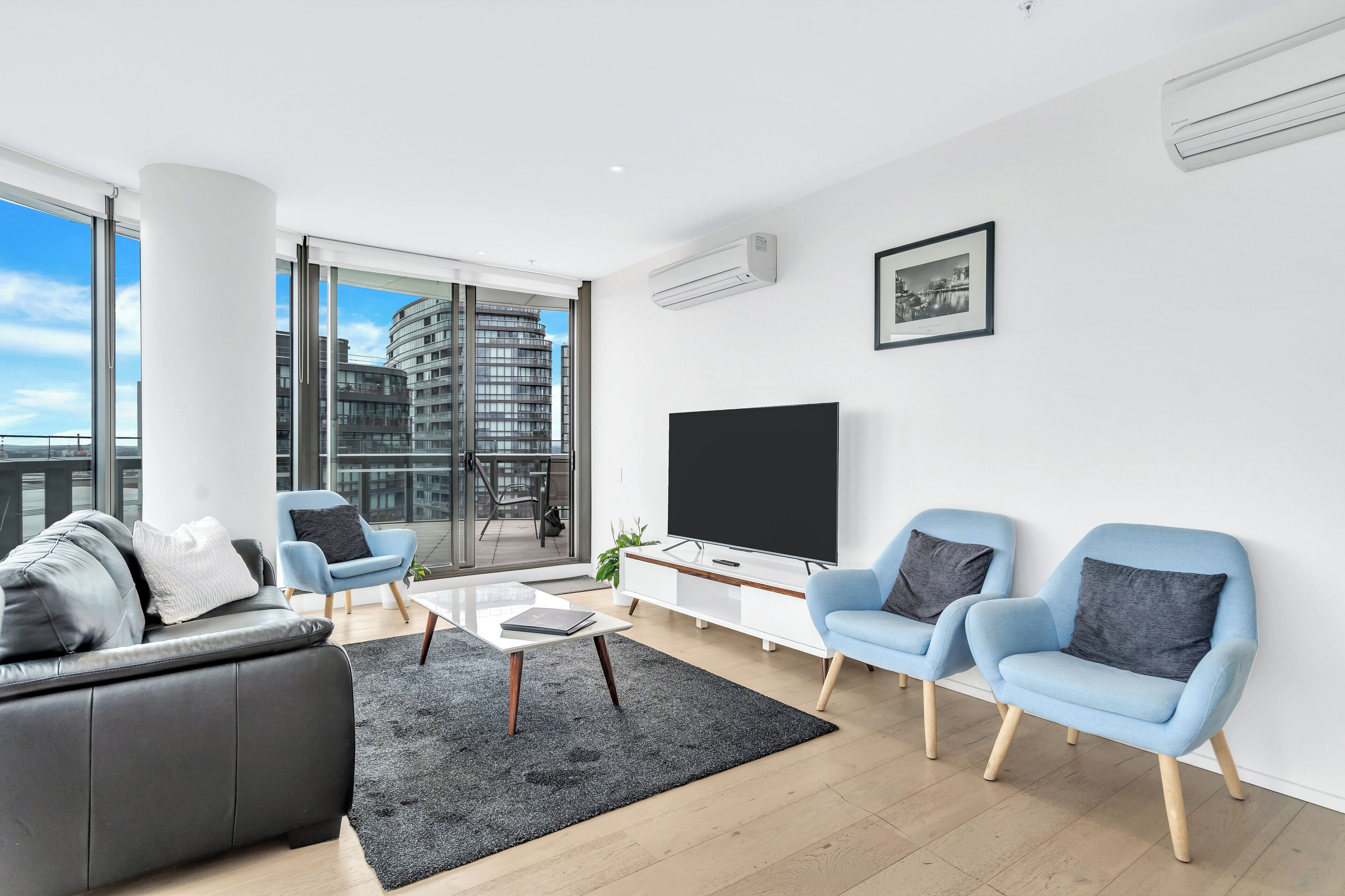 A modern living room with a black leather sofa, light blue armchairs, a glass coffee table, and a large flat-screen TV.