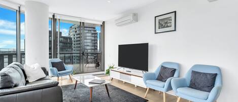 A modern living room with a black leather sofa, light blue armchairs, a glass coffee table, and a large flat-screen TV.