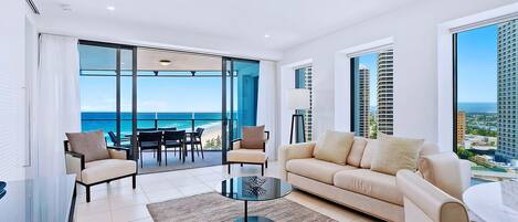 Una sala de estar moderna con un sofá, sillas y una mesa de centro. Se ve un ventanal con vistas al océano y edificios.