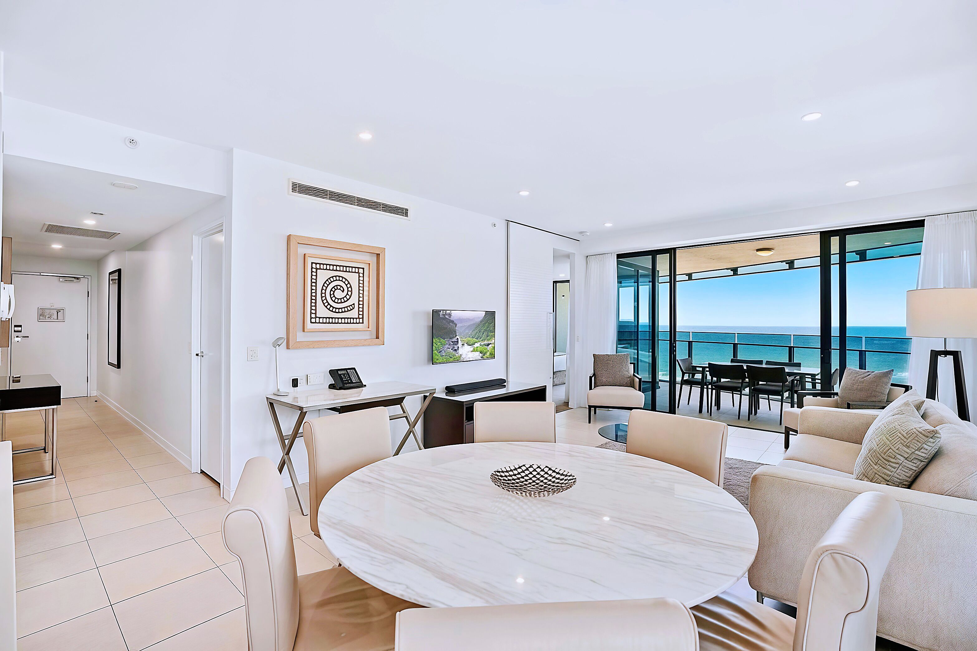 A modern living room with a marble table, beige sofas, and a large window overlooking the ocean.