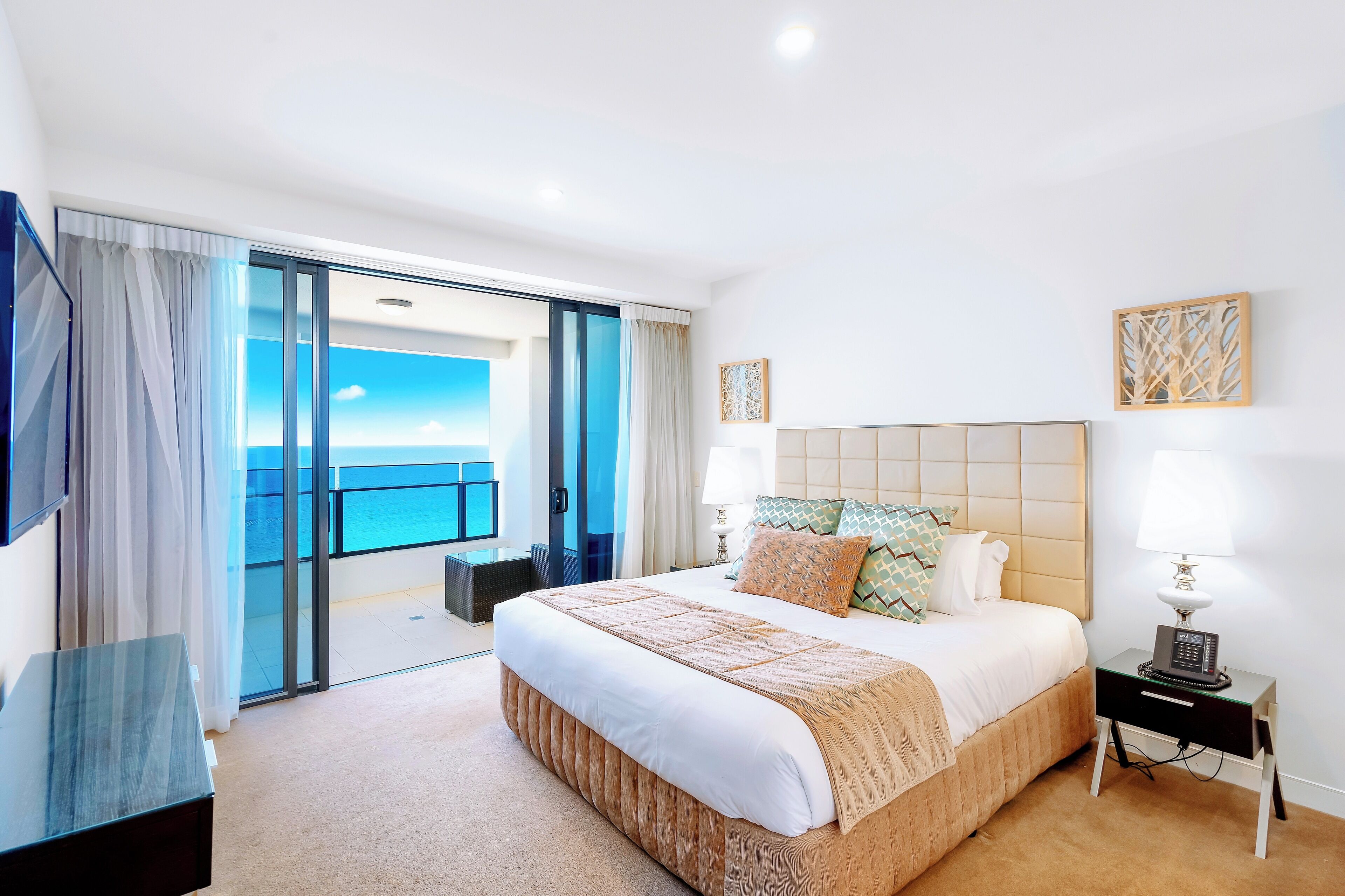 A modern hotel room with a large bed, a bedside table with a lamp, a view of the sea through a sliding door, and a small seating area.
