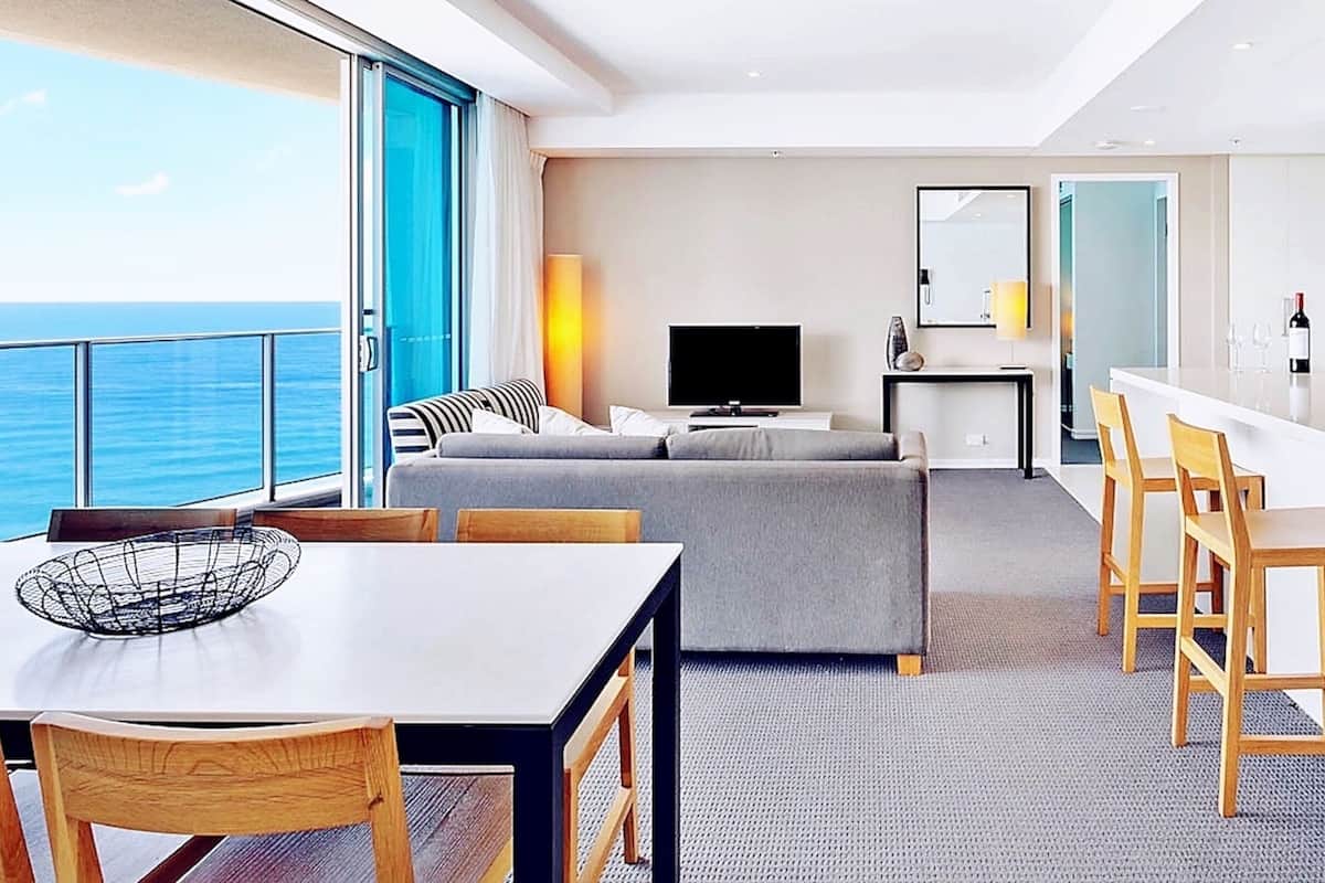 Een moderne, goed verlichte kamer met een groot raam met uitzicht op zee, een zithoek met een tafel en stoelen, een bank, een televisie en een klein keukentje.
