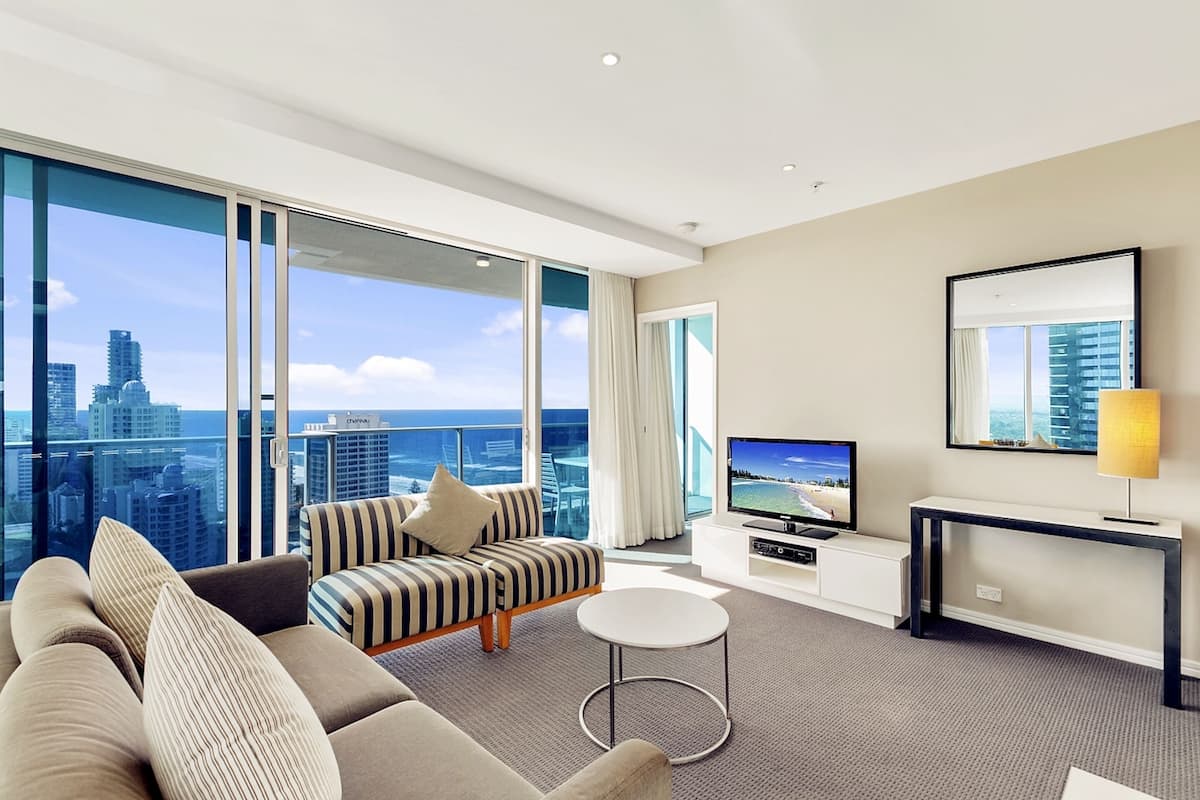 Een moderne hotelkamer met een groot raam met uitzicht over de stad, een comfortabele bank, een klein rond tafeltje, een televisie en een console met een lamp.