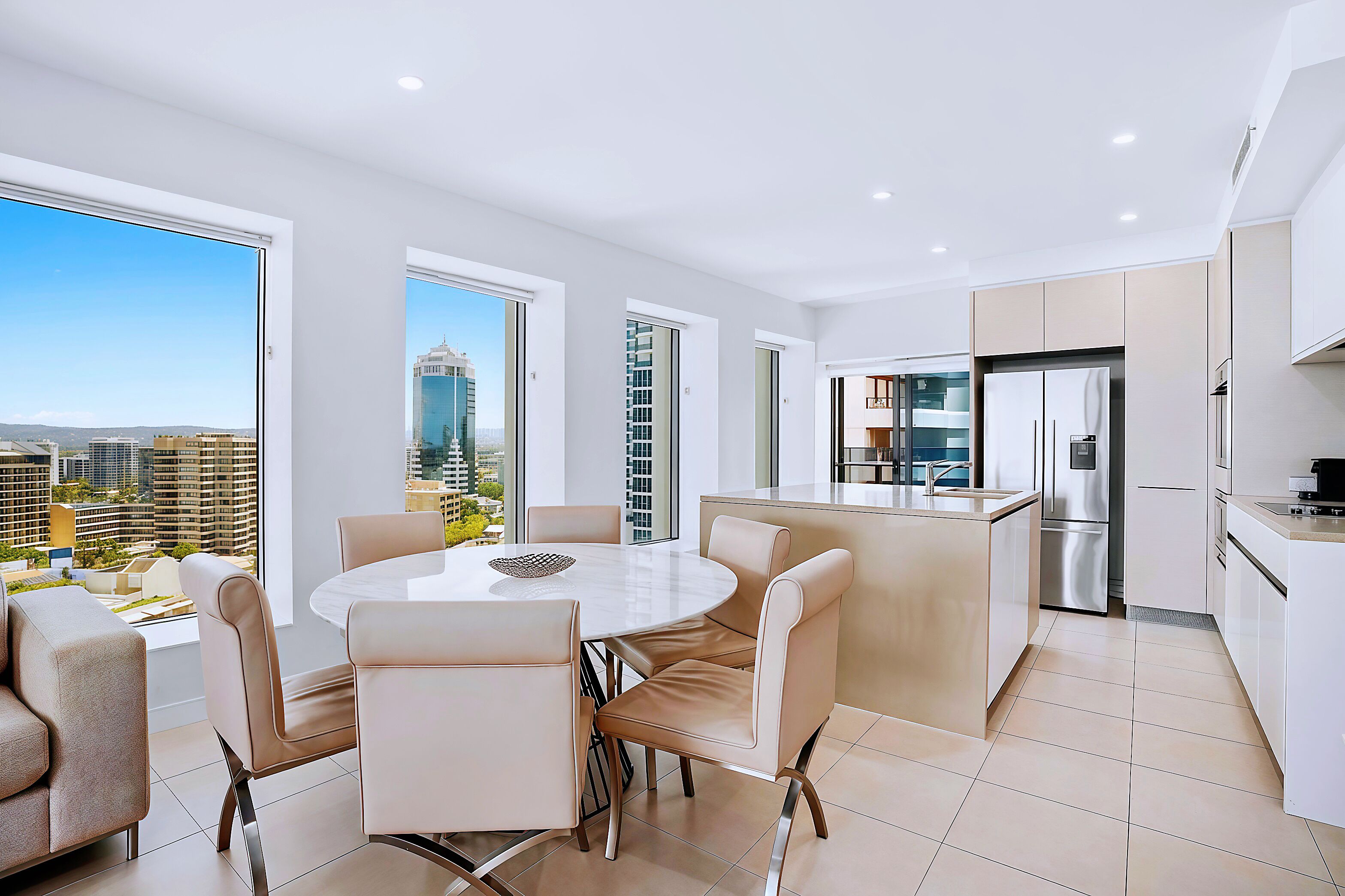 A modern kitchen with a central island, a dining area with a round table and chairs, and large windows offering a city view.