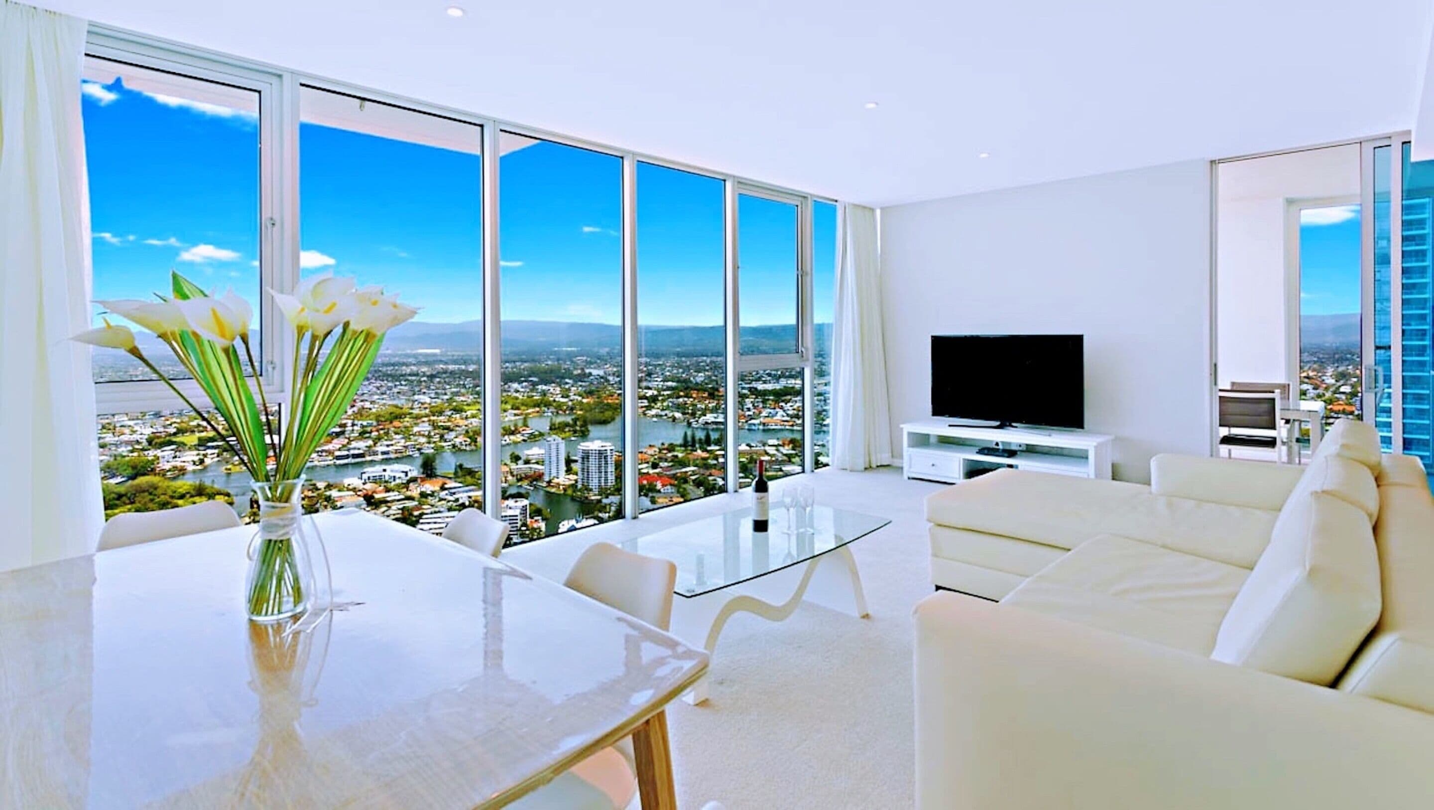 Ein modernes Wohnzimmer mit großem Fenster und Blick auf die Stadt, einem Flachbildfernseher sowie einem Essbereich mit einem Blumenstrauß.