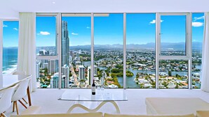 A modern hotel room with a large panoramic window, a dining area with a table and chairs, and a bottle of wine on the table. - H Luxury Residence Apartments - Holiday Paradise (Surfers Paradise)