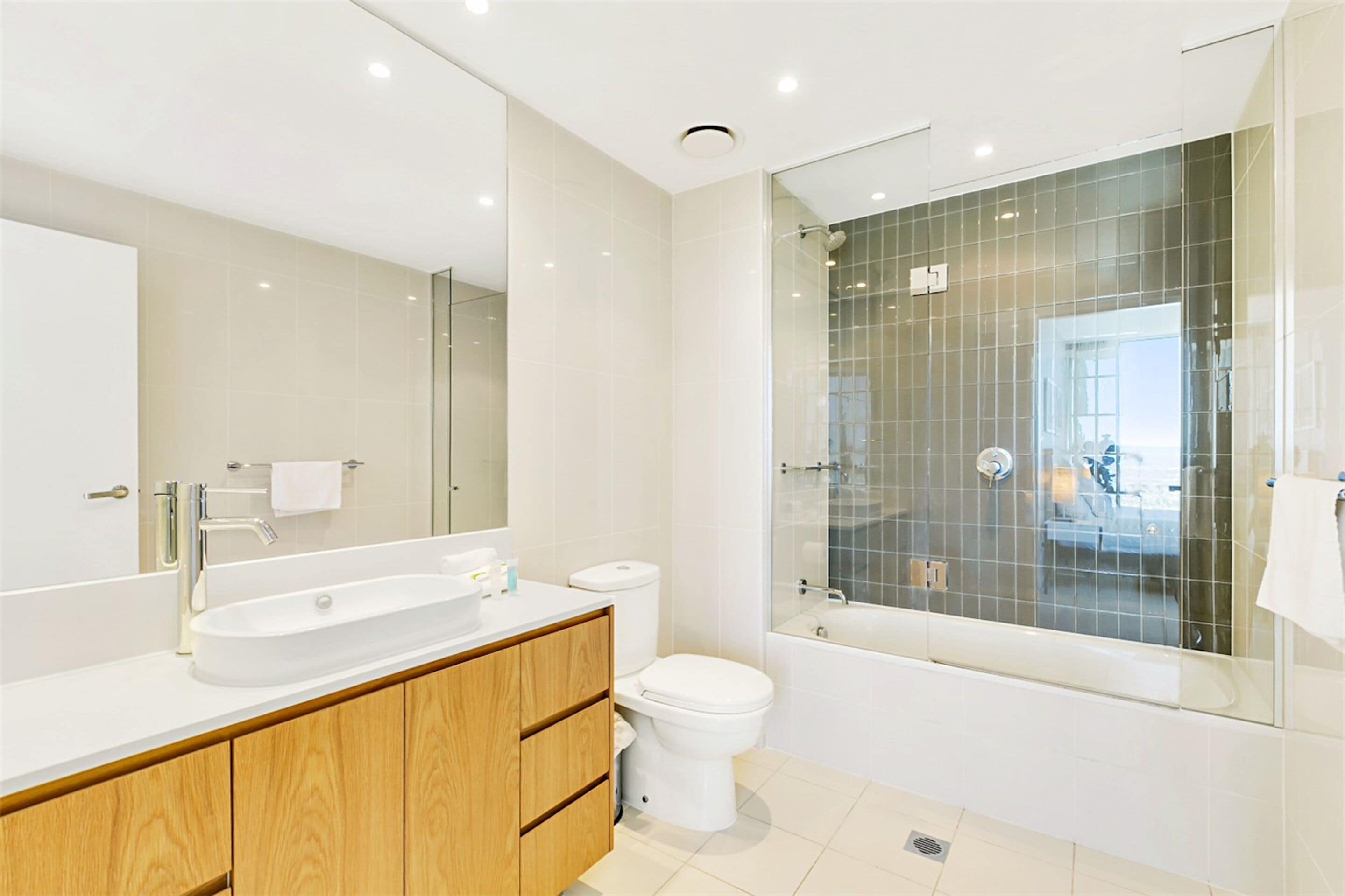 A modern bathroom with a large mirror, a wooden vanity, a white sink, a toilet, and a glass-enclosed shower area.