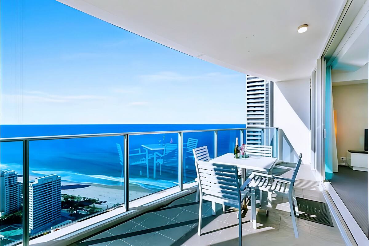 Een balkon met glazen hekwerk, een tafel met stoelen en uitzicht op de oceaan en gebouwen.