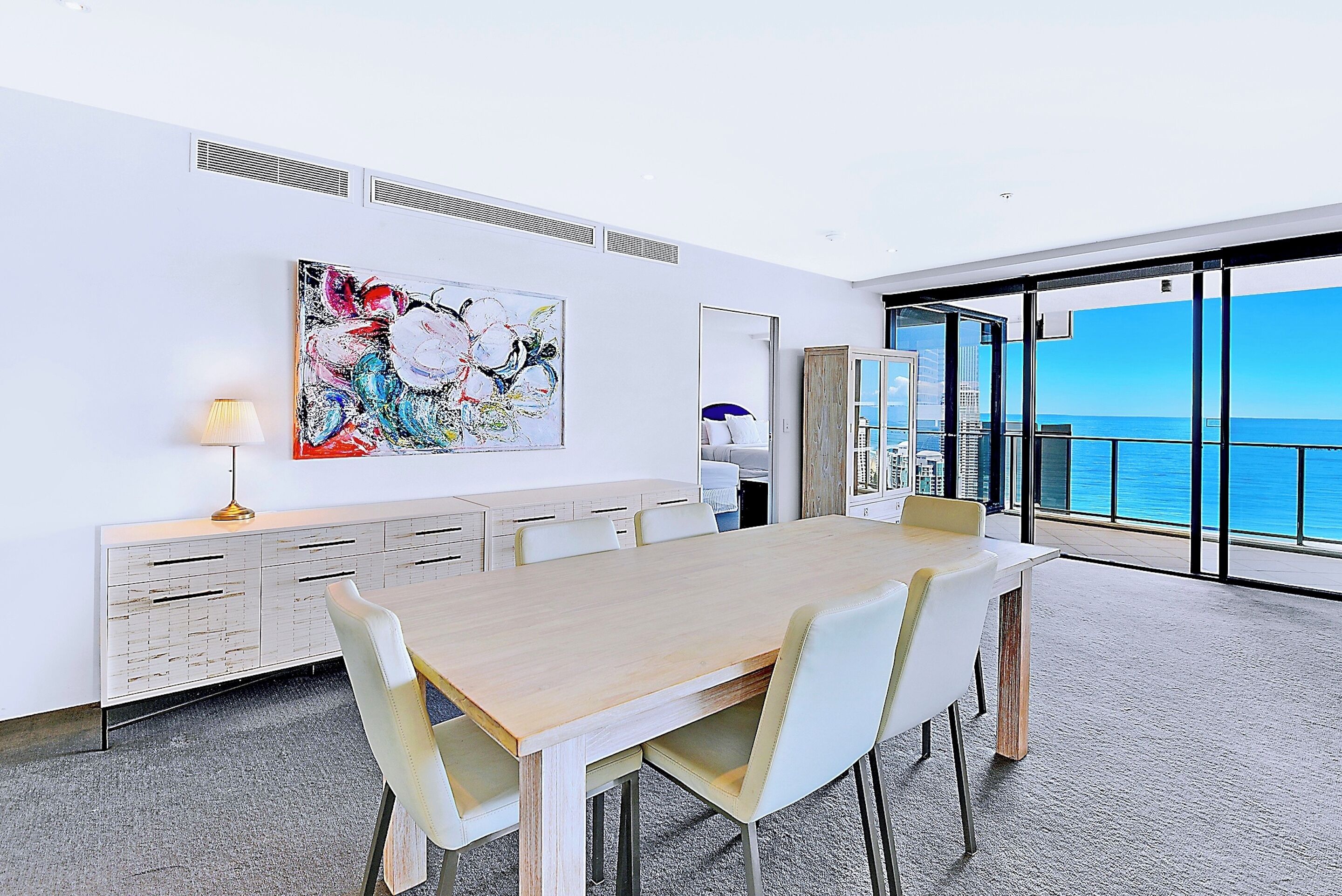 Quarto de hotel moderno com uma grande mesa de madeira, cadeiras brancas, um quadro na parede, um armário com gavetas e uma grande janela com vista para o oceano.