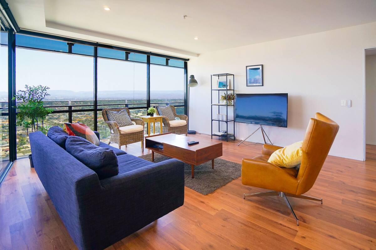 A modern living room with a large sofa, a coffee table, and a yellow armchair. There is a TV on the wall and a bookshelf.