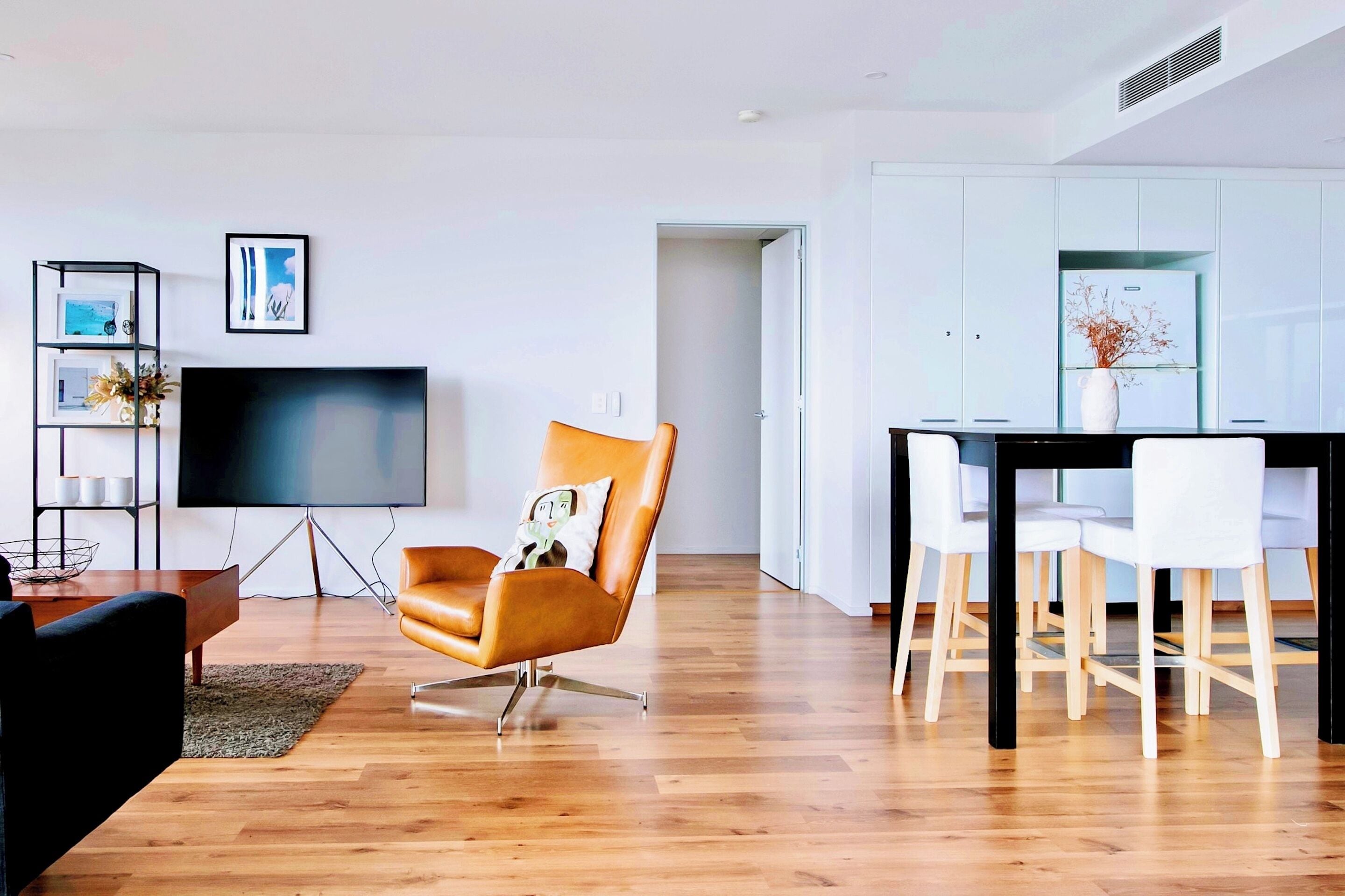 Uma sala de estar moderna com televisão de tela plana, uma poltrona de couro, uma mesa de jantar com cadeiras e um armário branco.
