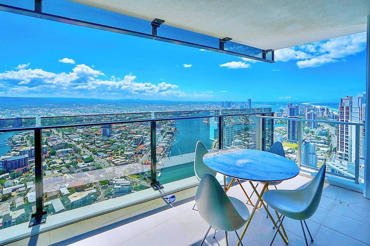 A high-rise balcony with a glass railing, a round table, and two chairs, overlooking a cityscape and a body of water.