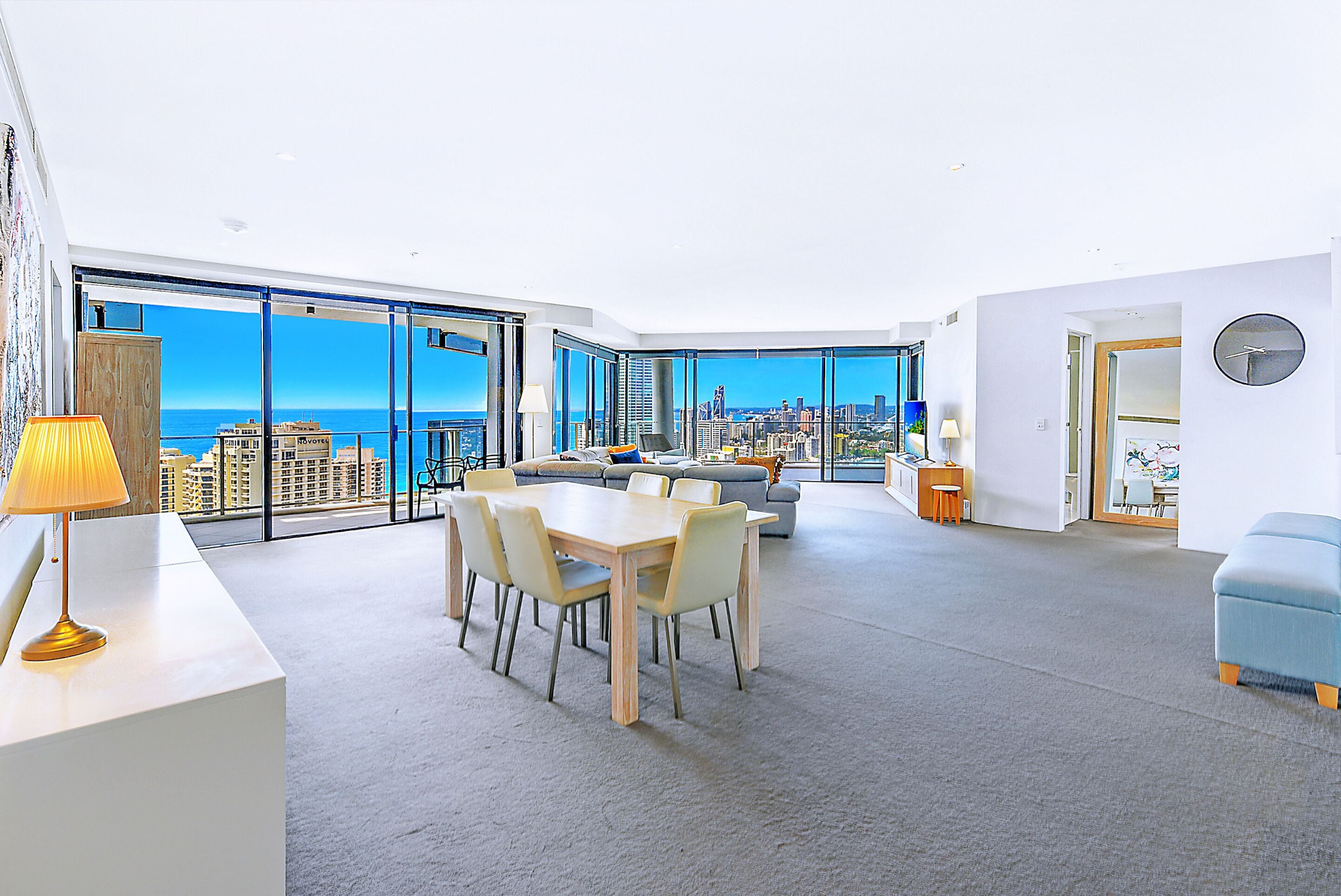 A spacious living area with a large dining table, a sofa, and a view of the cityscape through floor-to-ceiling windows.