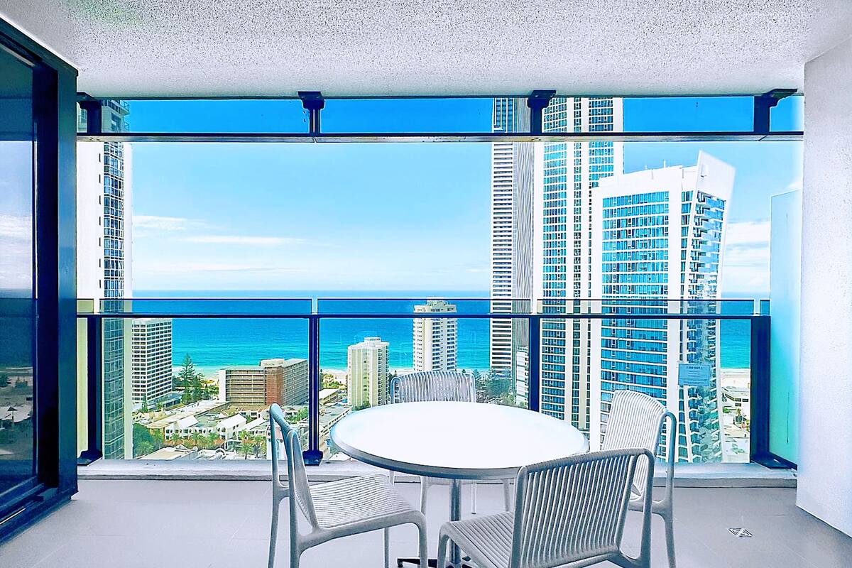 A balcony with a round table and two chairs, overlooking a cityscape with tall buildings and a view of the ocean.
