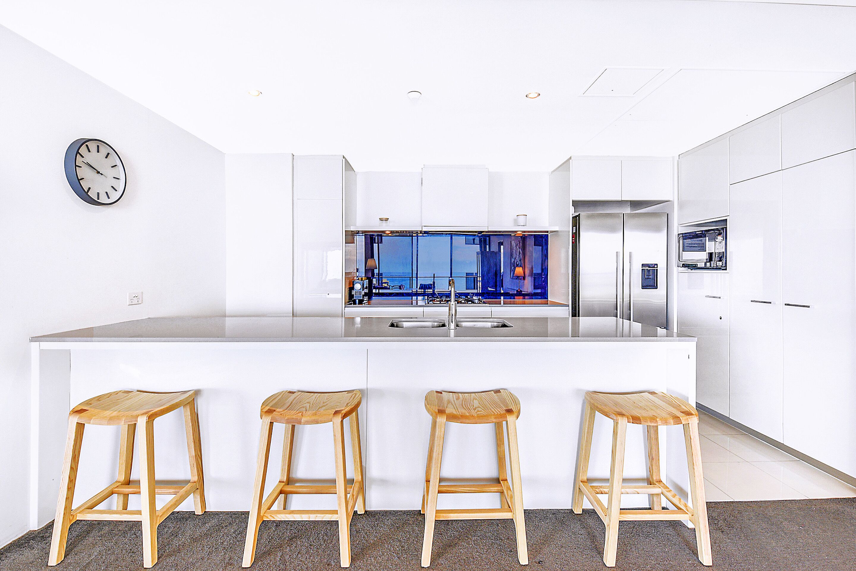 A modern kitchen with a large island, bar stools, a clock, and a view of the city through the windows.