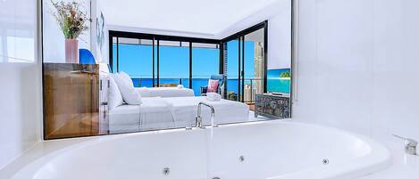 A modern bathroom with a large white bathtub, a glass-enclosed shower area, and a view of the sea through large windows.