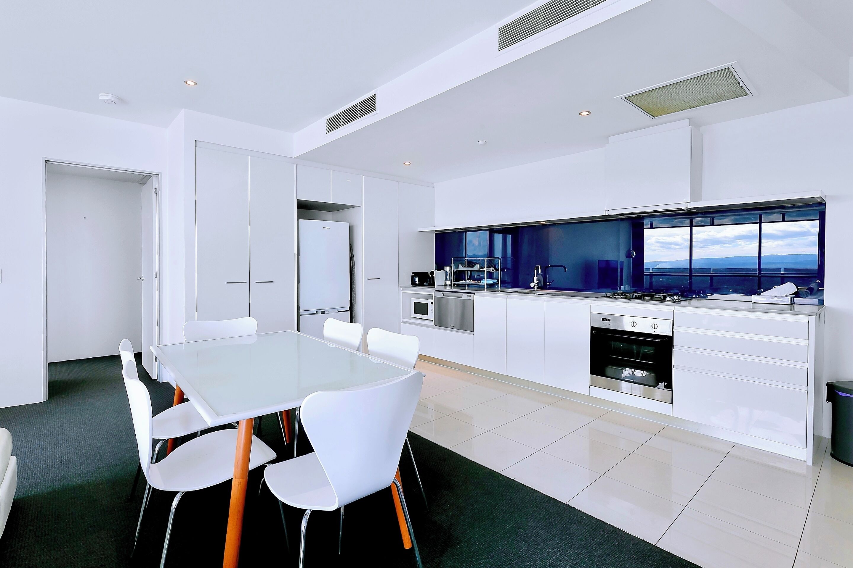 A modern kitchen with white cabinetry, a large island with a built-in oven, and a view of the city through large windows.