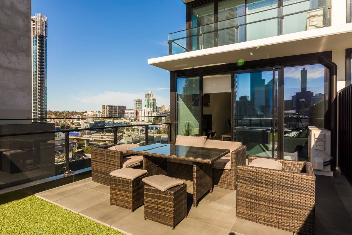 A modern outdoor patio with wicker furniture, a glass-top table, and a city view.