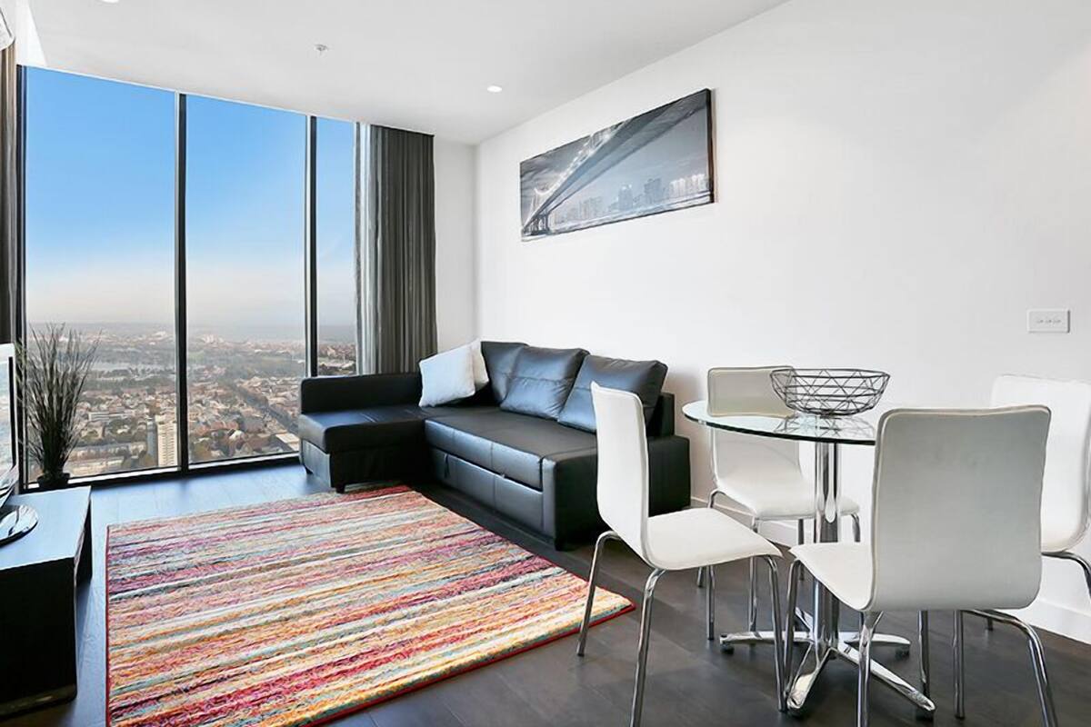 A modern living room with a black leather sofa, white chairs, a glass dining table, and a colorful striped rug.