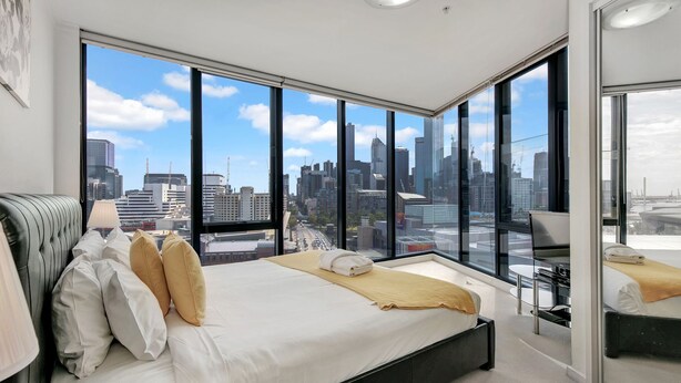 A modern hotel room with a large bed, a desk, and a view of a city skyline through floor-to-ceiling windows. - MJ Shortstay Whiteman St Apartments (Southbank)