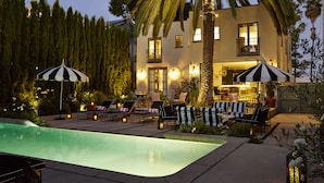 Un espace aménagé au bord de la piscine, avec des chaises longues et des parasols, une piscine, et un bâtiment doté de nombreuses fenêtres et de balcons.