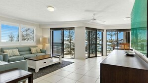 A spacious living room with a sofa, coffee table, and a dining area with a table and chairs. There is a large window with a view of trees and a balcony.