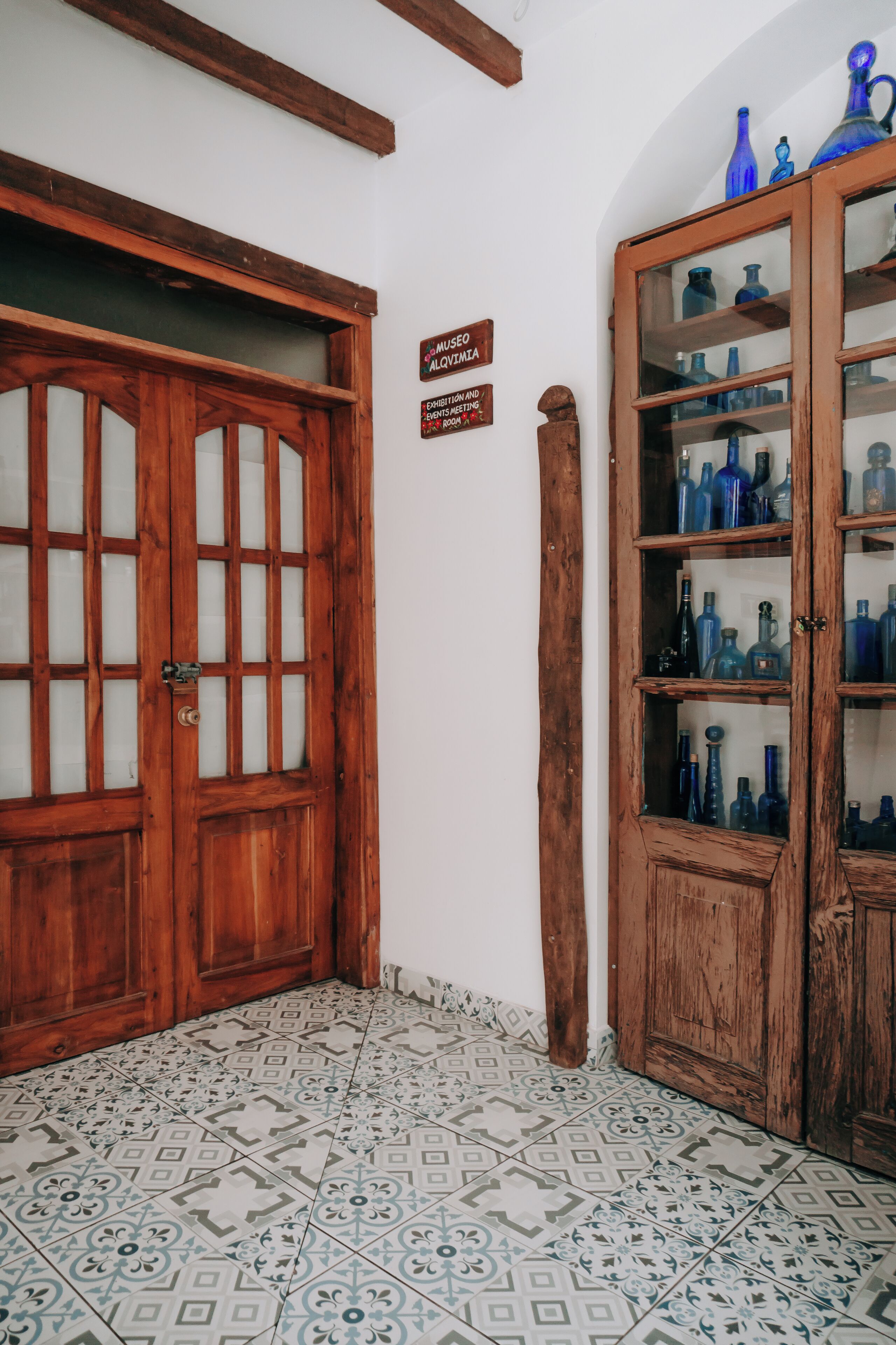 A room with a wooden door, a cabinet with blue glassware, patterned floor tiles, and a sign indicating a museum exhibit.