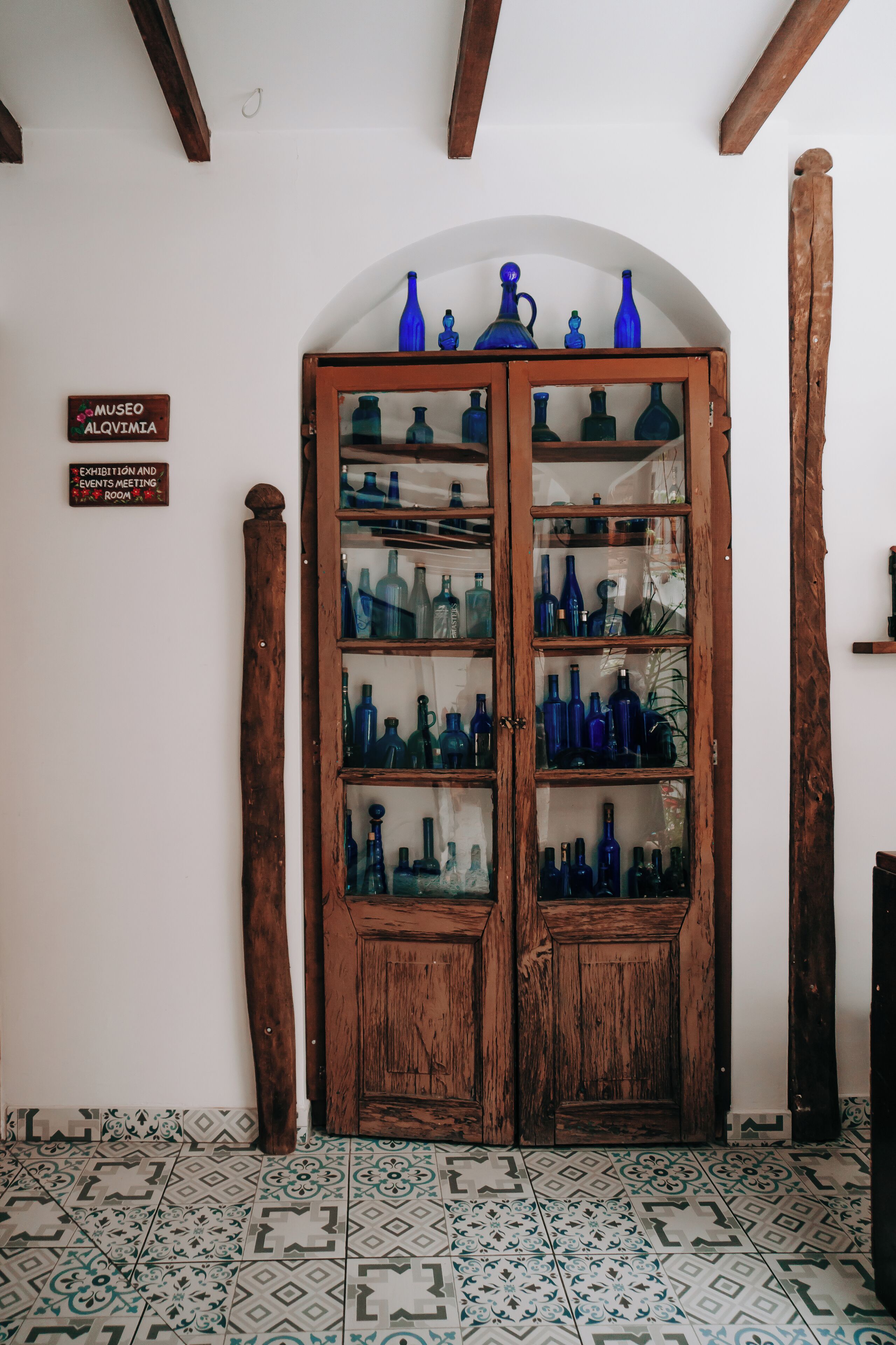 Una puerta de madera con paneles de vidrio que muestran botellas azules, un cartel que indica una sala de exposición de museo y baldosas de suelo con diseños.