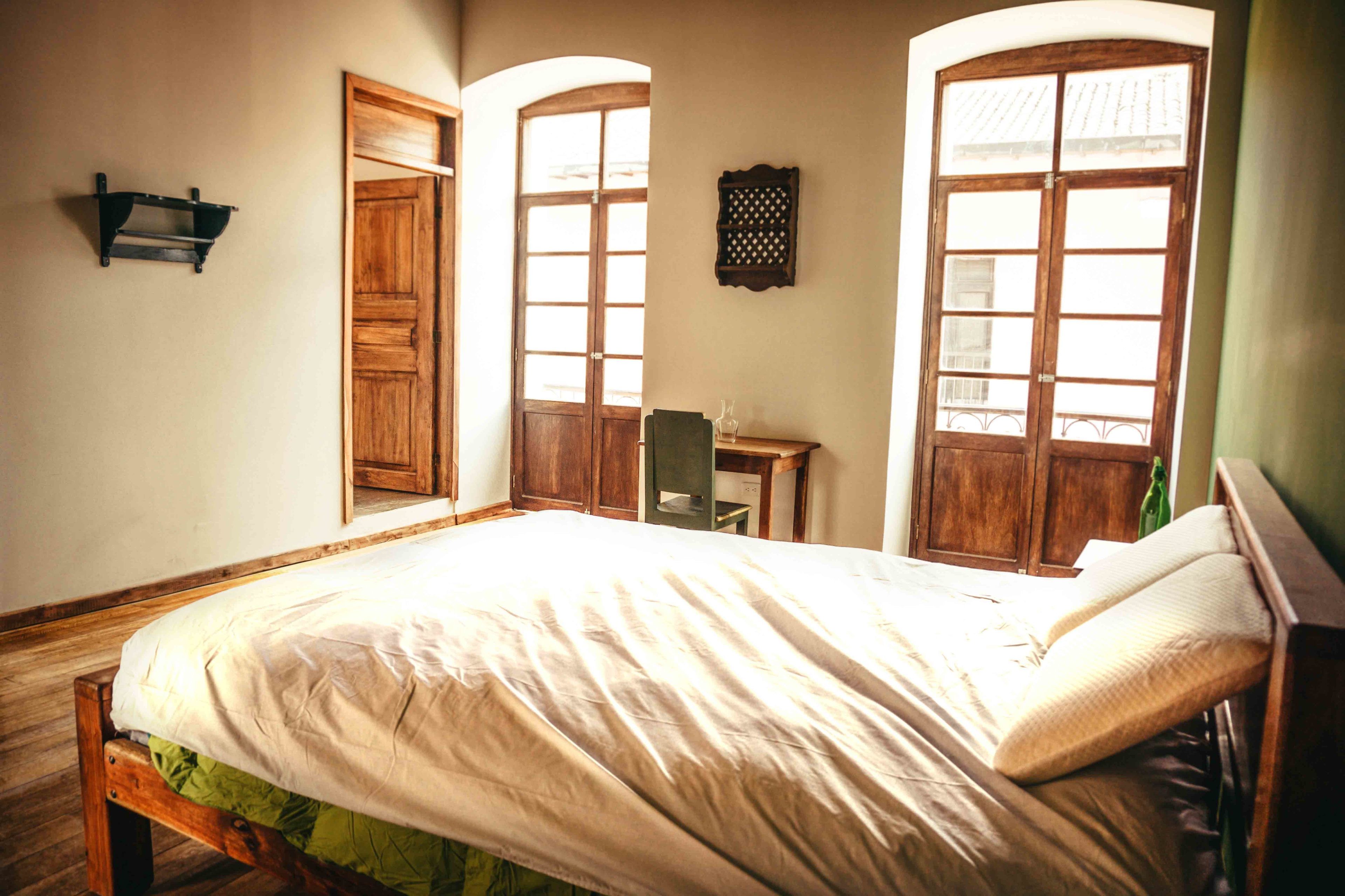 A bedroom with a bed, wooden wardrobe, arched windows, and a small desk.