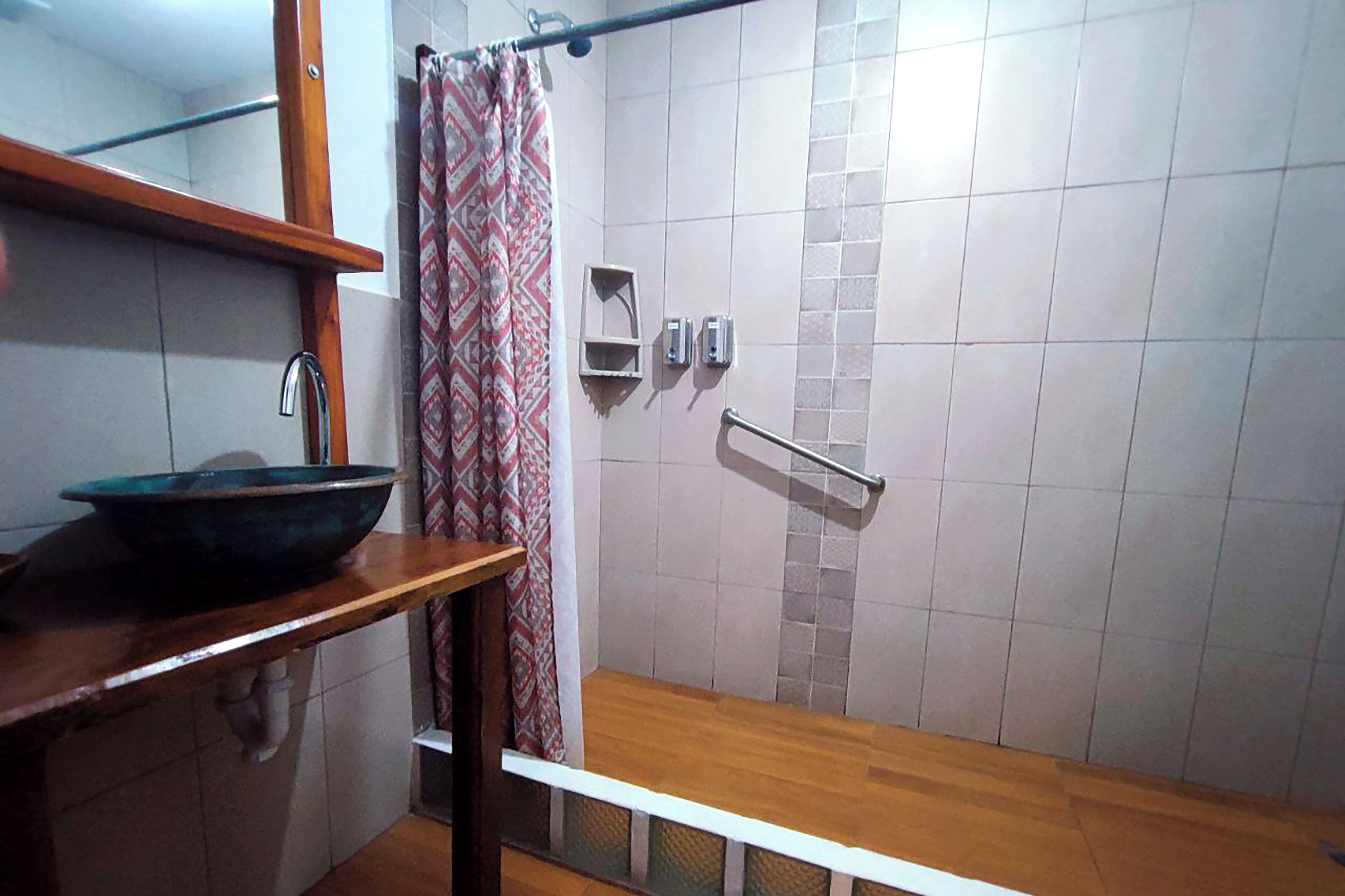 Un baño con tocador de madera, un lavabo grande, ducha con barra de apoyo metálica y cortina de ducha estampada.