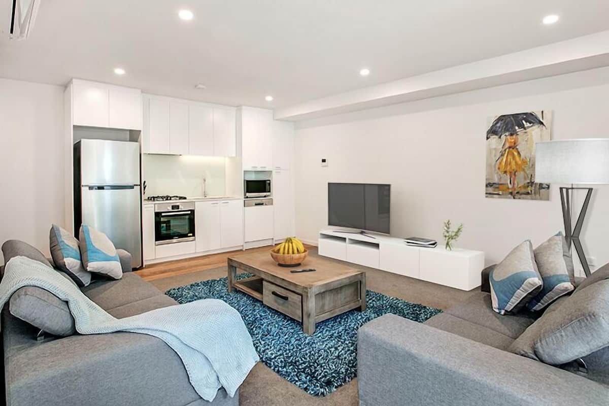 A modern living room with a sofa, coffee table, and TV. There is a kitchen area with white cabinets and appliances.
