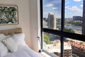 Un dormitorio con un ventanal que ofrece vistas a la ciudad, una cama con ropa de cama blanca y una planta enmarcada en la pared.