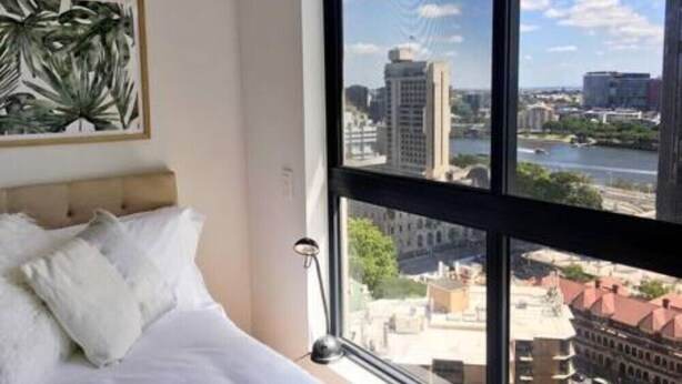 Ein Schlafzimmer mit großem Fenster und Blick auf die Stadt, einem Bett mit weißen Leinen und einer gerahmten Pflanze an der Wand.