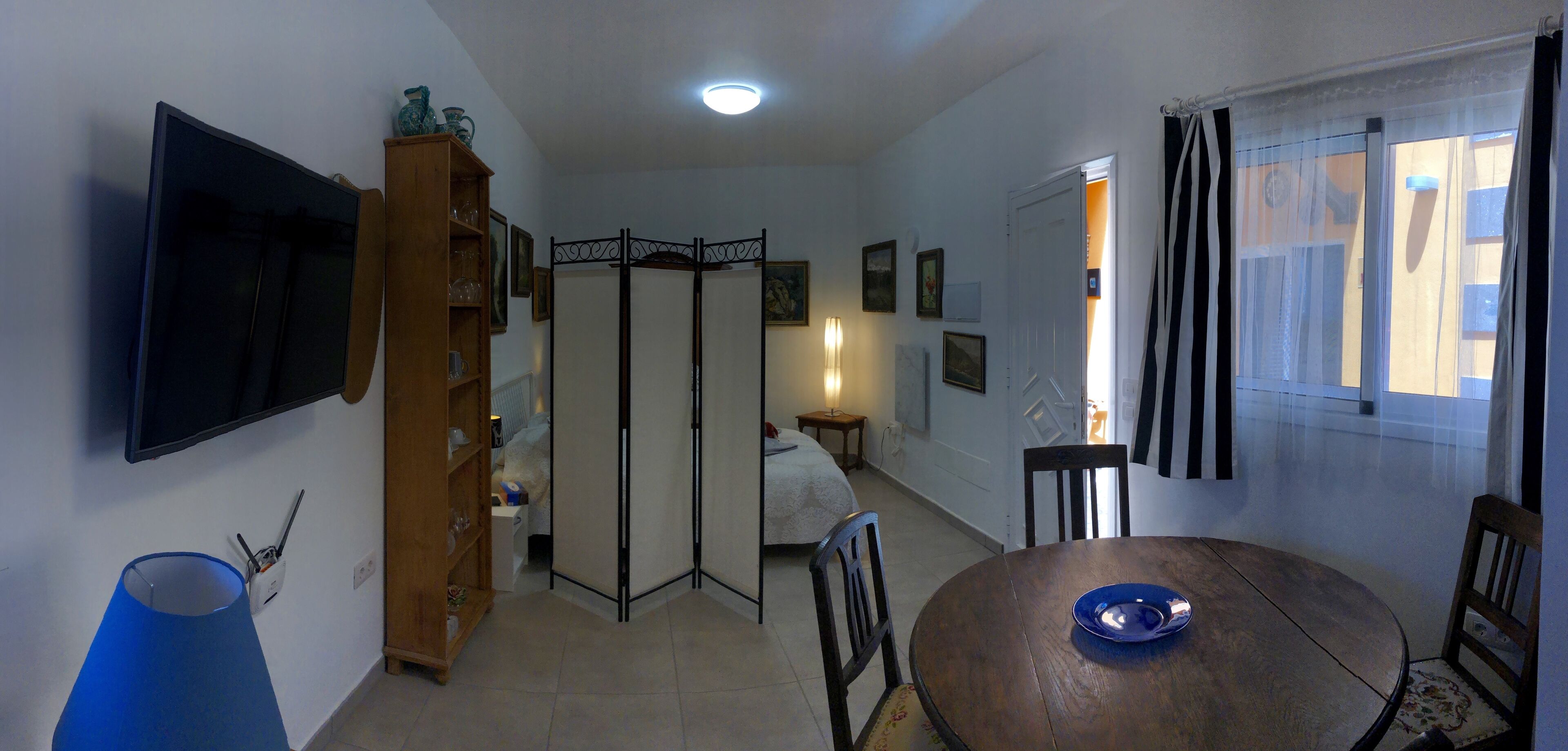 A room with a TV mounted on the wall, a dining table with chairs, a bookshelf, a partition, and a window with curtains.