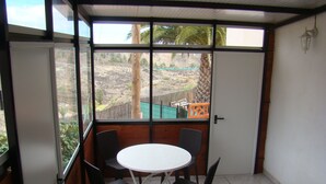 Un balcon avec une table et quatre chaises, une porte et une vue sur un espace clos, agrémenté d’arbres et d’une colline.