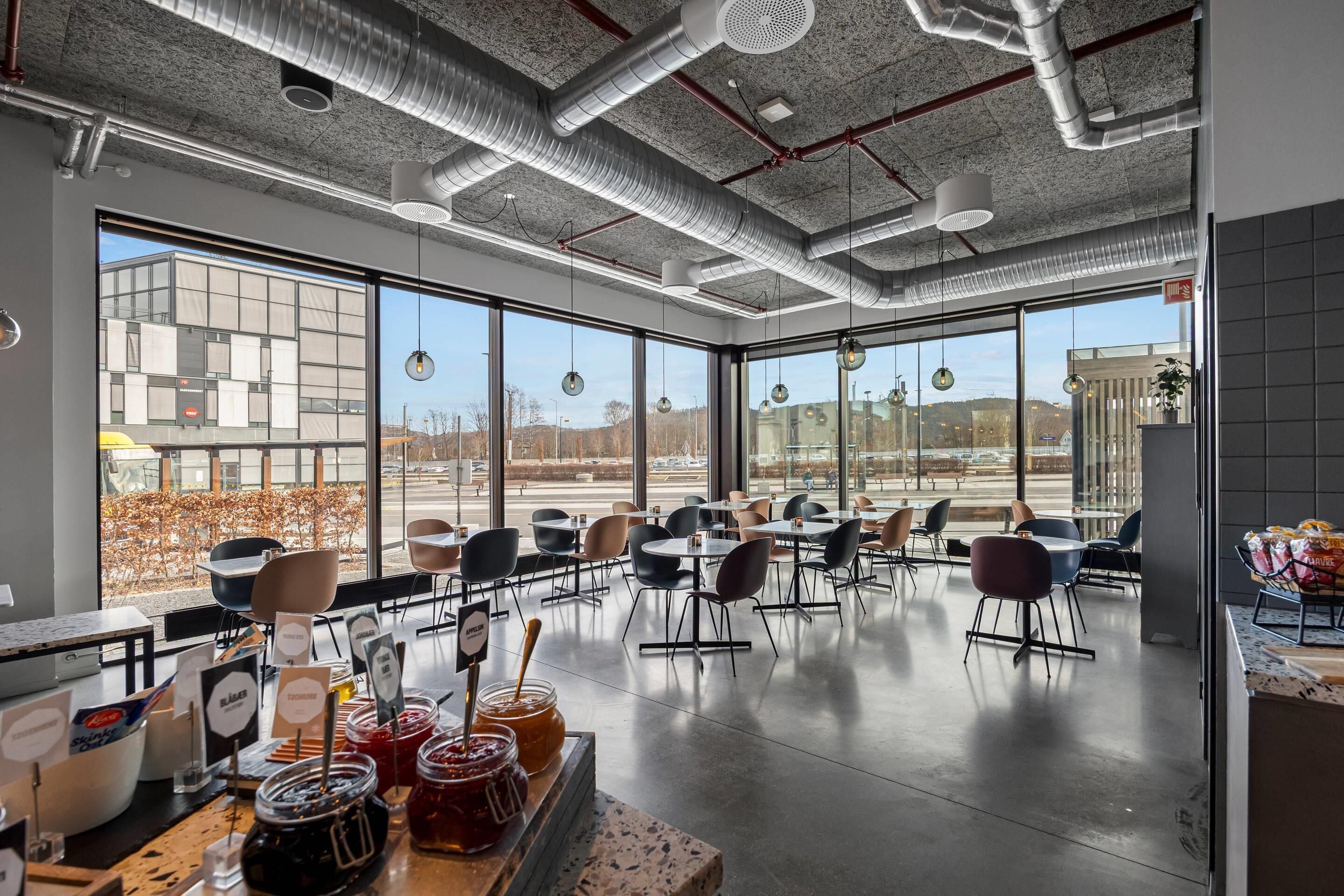 Een modern café met grote ramen, openliggende ventilatiekanalen en diverse zithoeken.