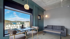 A modern room with a dining table, chairs, a sofa, and a large window offering a view of buildings and hills.