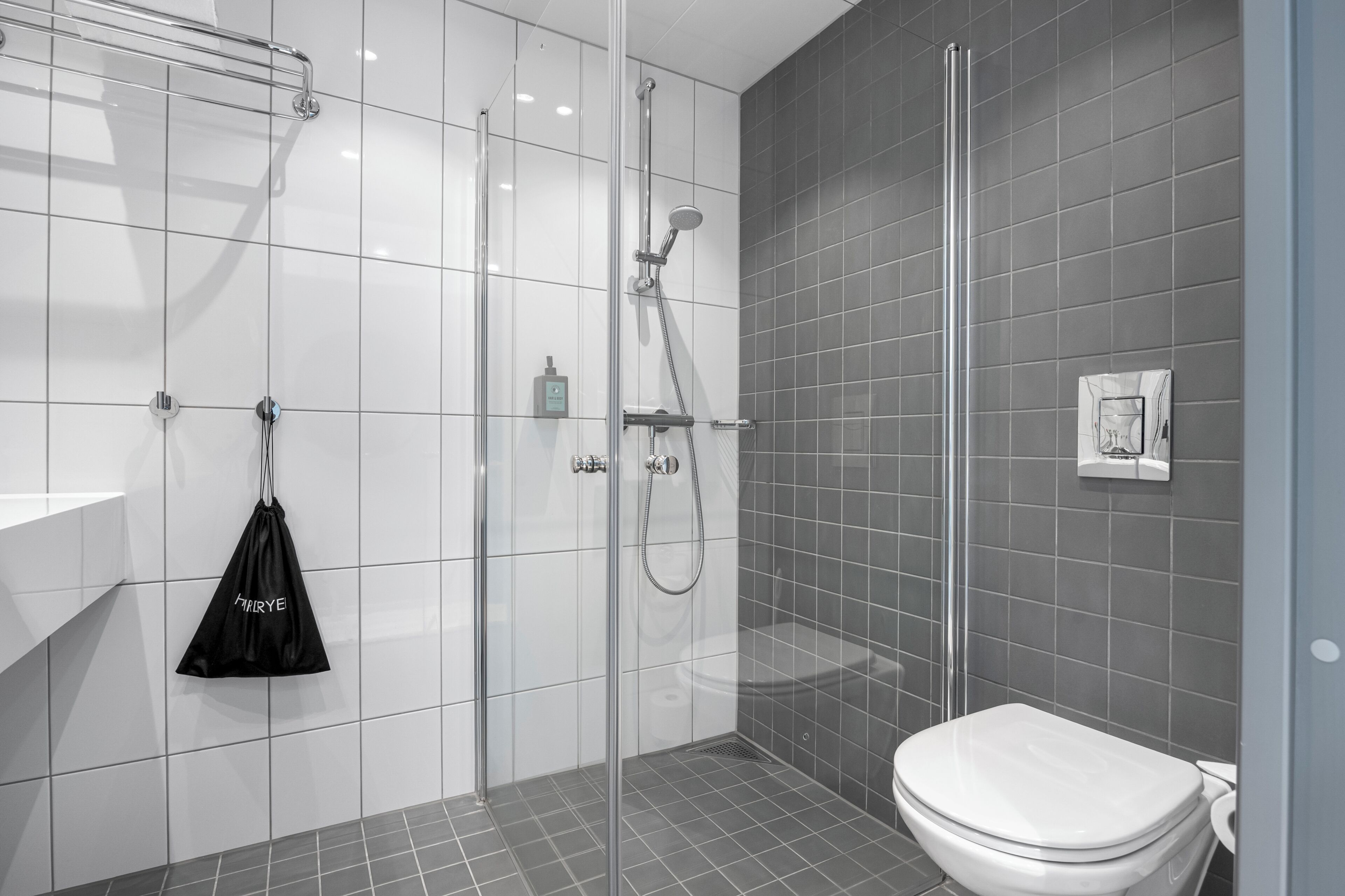 Un baño moderno con cabina de ducha de vidrio, inodoro blanco y un bolso negro colgado en un perchero.