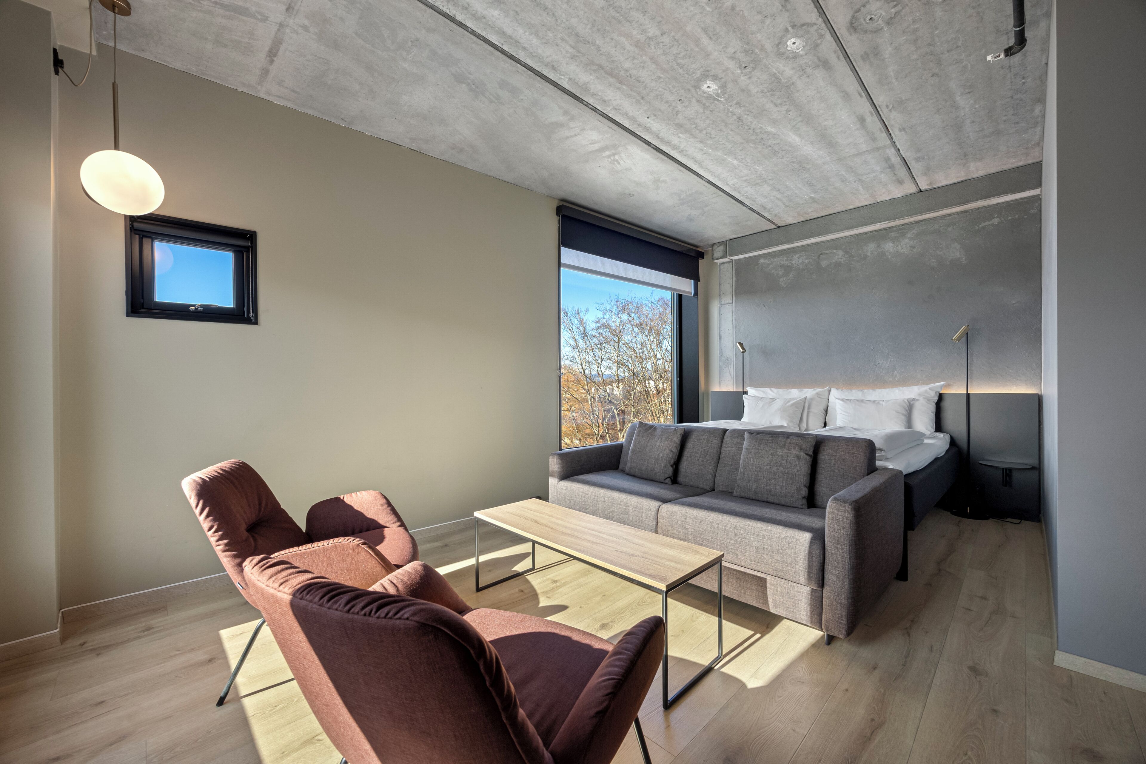 A modern room with a sofa, two chairs, a small table, a window with a view, and a bed with a headboard.