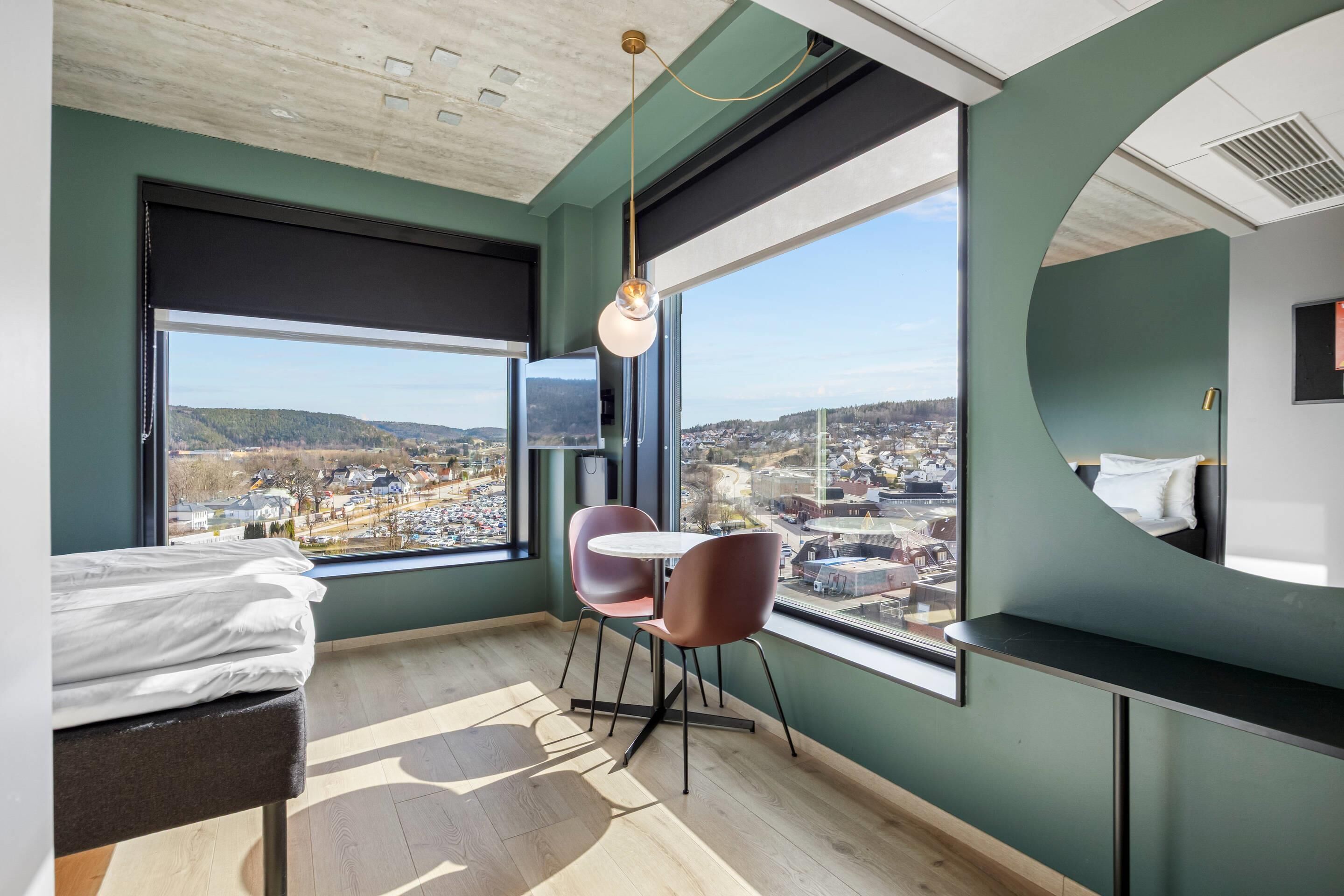 A modern hotel room with a large window, a bed, a round table, and a chair.