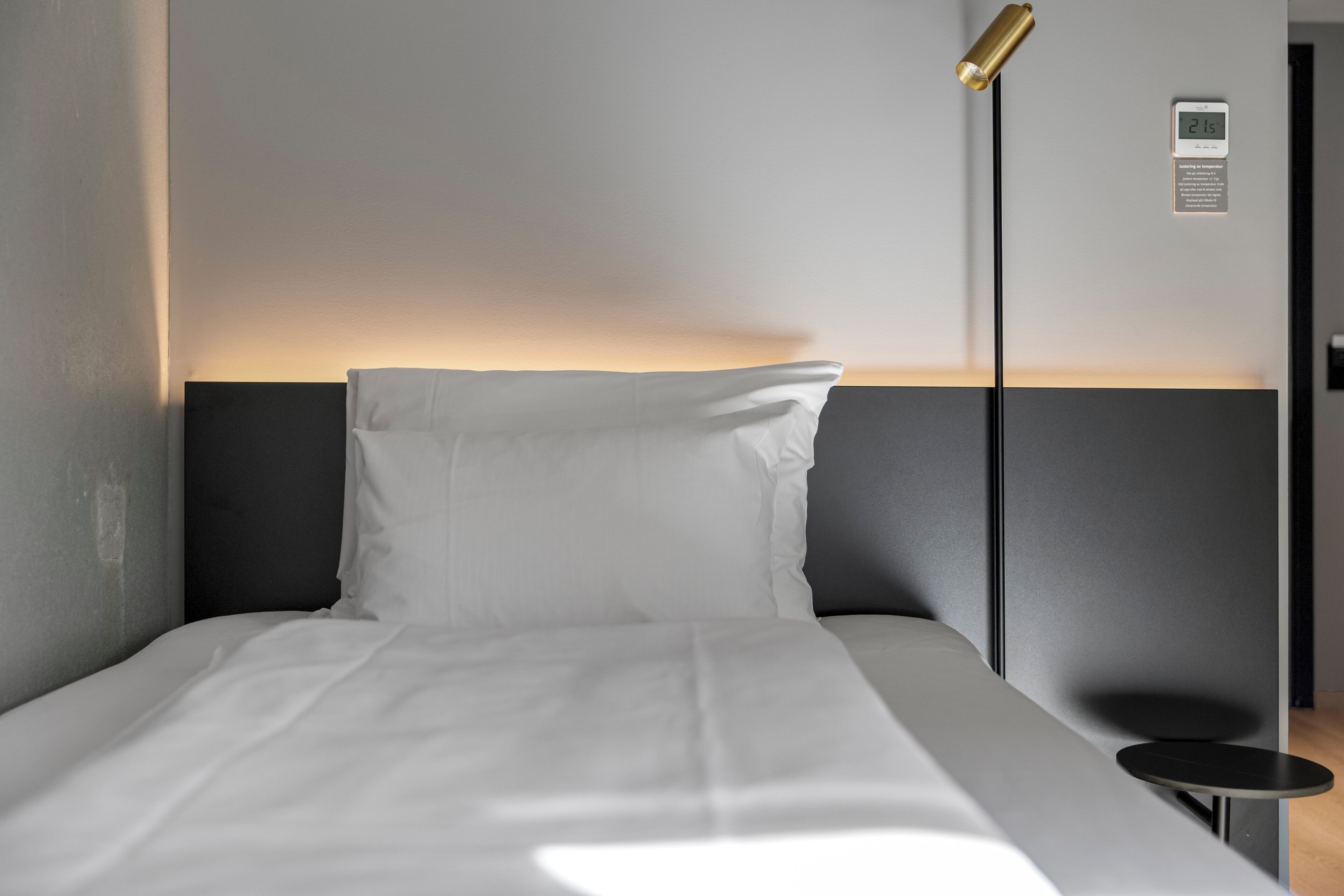 A neatly made bed with white linens, a bedside table, a modern floor lamp, and a digital clock on the wall.