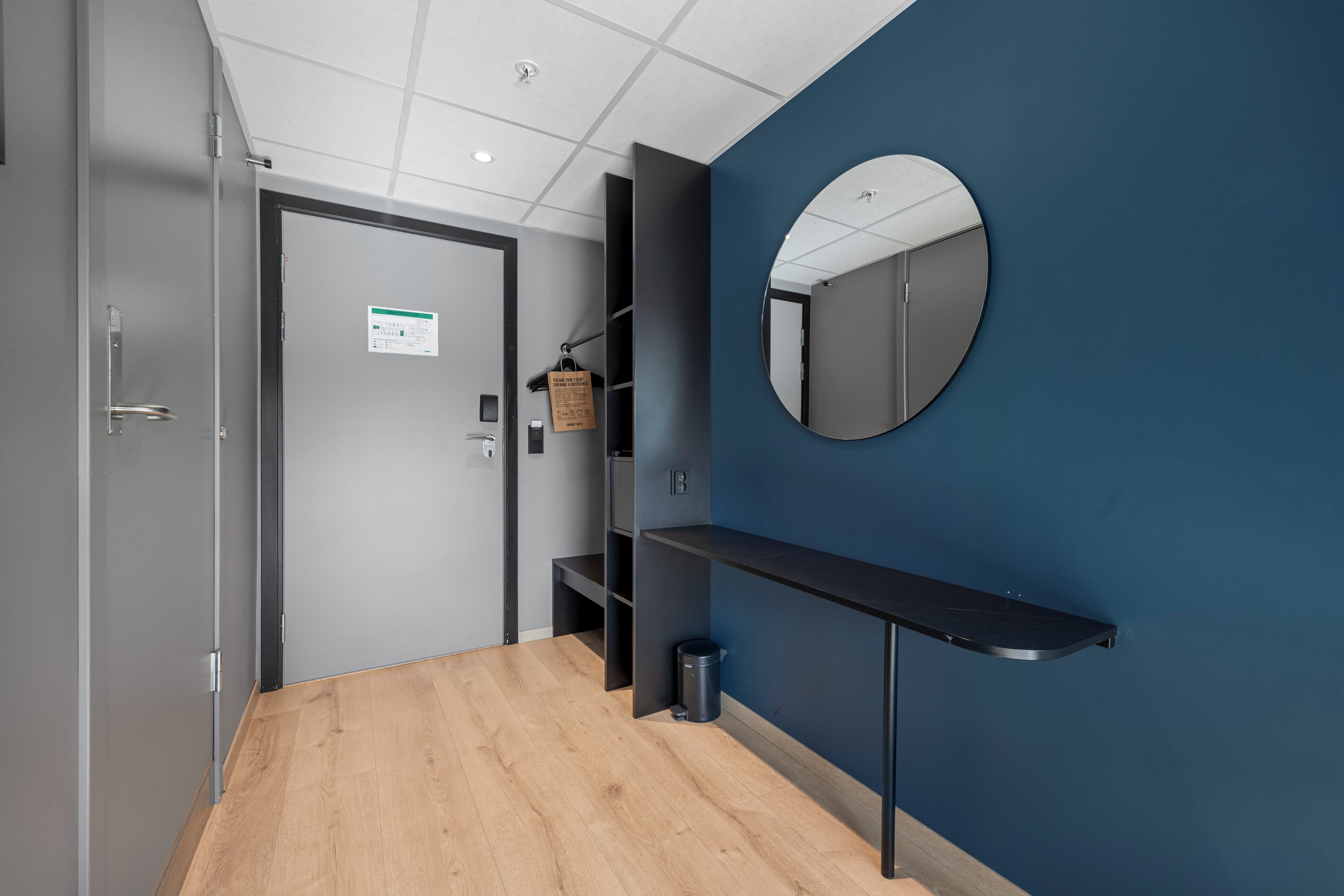Un pasillo con suelo de madera, pared azul, espejo redondo, estante negro y una puerta con un letrero.