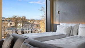 A bedroom with a large window, a bed with white linens, and a view of buildings and trees.