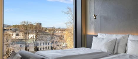 A bedroom with a large window, a bed with white linens, and a view of buildings and trees.