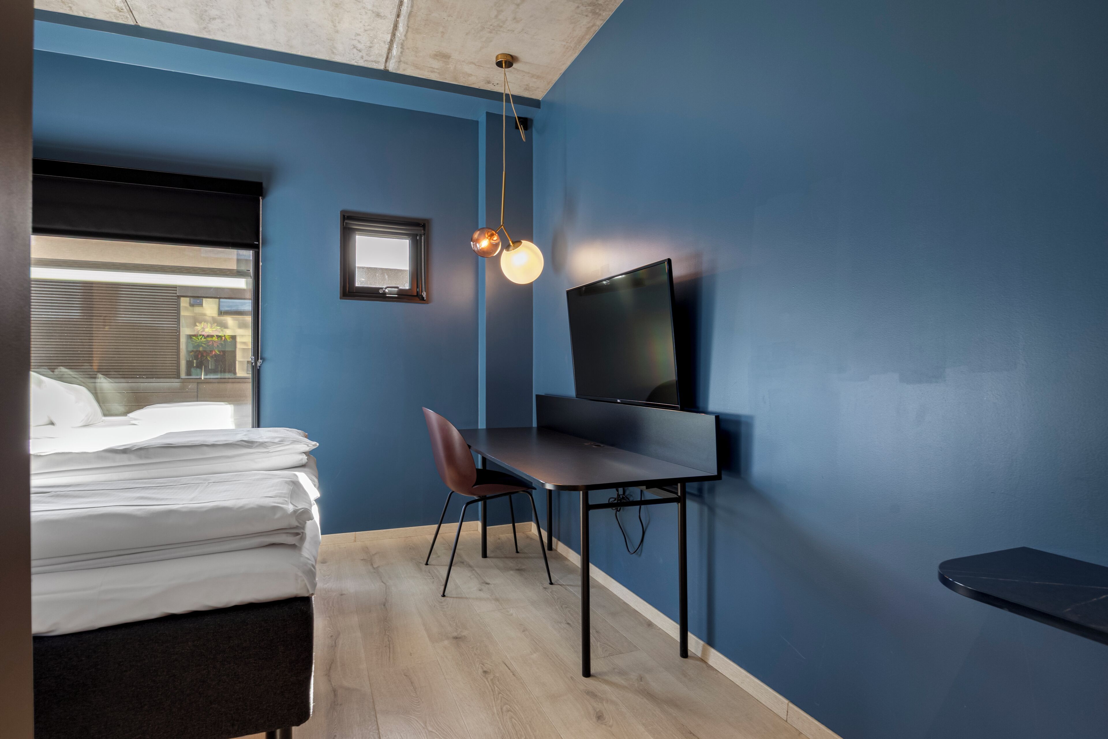 Une chambre avec un lit, un bureau et une chaise, une télévision et une fenêtre avec des stores.